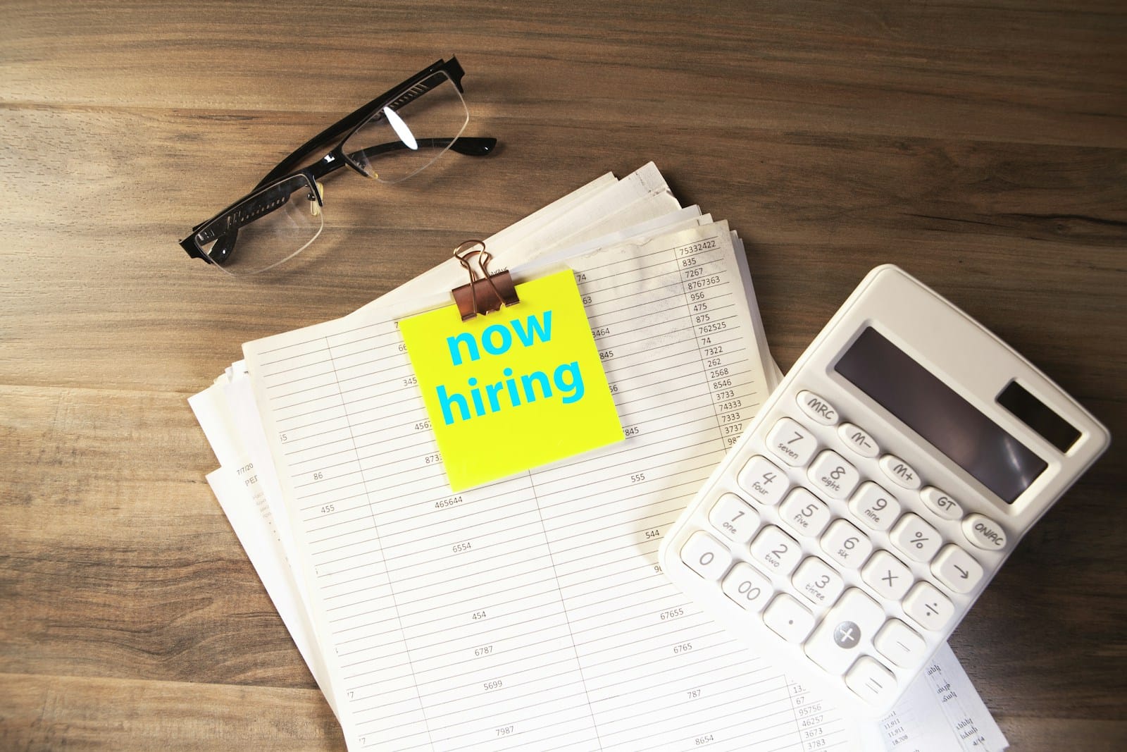 電卓と眼鏡、書類にクリップで留めた「now hiring」のメモ